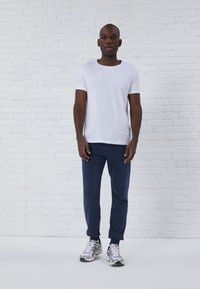 Un t-shirt blanc à manches courtes, un pantalon de survêtement bleu marine et des chaussures de sport argentes avec des accents foncés sont portés par une personne devant un fond en briques blanches.