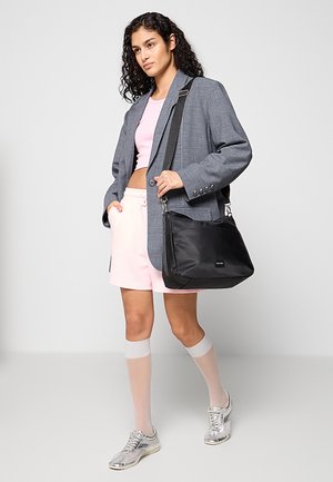 Černá nylonová taška přes rameno s nastavitelným popruhem; má hladký povrch a malé logo; nese ji osoba v šedém saku a růžových šortkách.