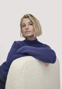 Königsblauer Strickpullover mit hohem Ausschnitt und strukturierten Ärmeln, getragen von einer Person, die auf einem cremefarbenen, strukturierten Kissen sitzt.