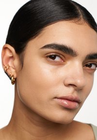Närbild av ett ansikte med slät hy, mörkt hår och framträdande drag. Gyllene hoop örhängen, naturlig läppfärg och välskötta ögonbryn.