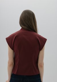Bordowy top z krótkim rękawem z kołnierzykiem i wyraźnie zarysowanymi ramionami, o dopasowanym kroju i gładkiej tkaninie. Widok z tyłu pokazuje detal szycia.