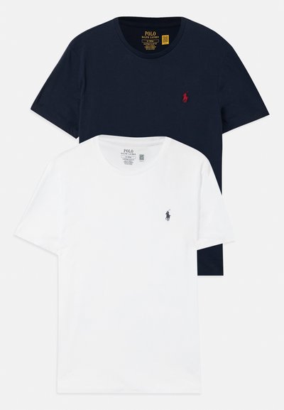 Dve crew-neck Polo Ralph Lauren majice, ena mornarsko modra z rdečim logotipom in ena bela s temnim logotipom, položeni na beli podlagi.