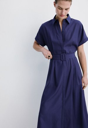 Femeie care ajustează centura unei rochii midi bleumarin cu mâneci scurte, nasturi și guler, pe un fundal luminos simplu.