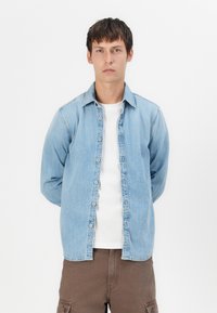 Lichtblauw denim overhemd met een knoopsluiting, lange mouwen en een puntige kraag, gedragen over een wit T-shirt. Achtergrond is volledig wit.