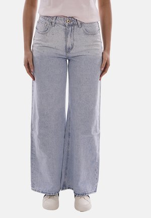 Persona che indossa jeans larghi azzurri con decorazioni di strass sulla parte superiore frontale, scarpe da ginnastica bianche e una camicia bianca infilata dentro.