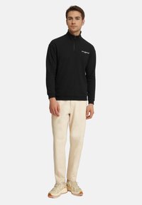 Pullover in pile nero con zip e colletto alto, dotato di un logo sul petto. Abbinato a pantaloni chiari e sneakers beige.