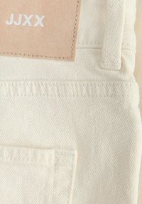 Zbliżenie kremowych spodni dżinsowych pokazujące tylną kieszeń, szlufkę na pasek, szczegóły przeszyć oraz beżową metkę z białym napisem "JJXX".