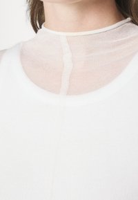White sleeveless top with a high, sheer neckline; features a smooth texture and a subtle vertical seam detail.
