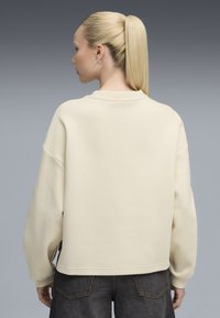 Sudadera de color beis claro con un corte relajado, mangas oversized y puños acanalados. Combinada con pantalones de mezclilla oscuros. Textura suave, detalles mínimos.