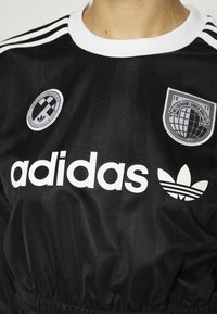 Svart Adidas-tröja med vit krage, Adidas-logotyp och två broderade lappar med geometriska mönster på bröstet.