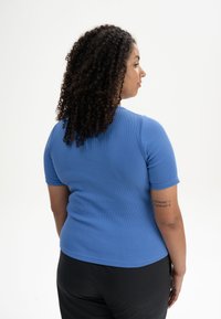Ribbed, short-sleeve top in bright blue, fitted design. Soft texture with a round neckline, worn with black pants. Visible tattoo on arm.