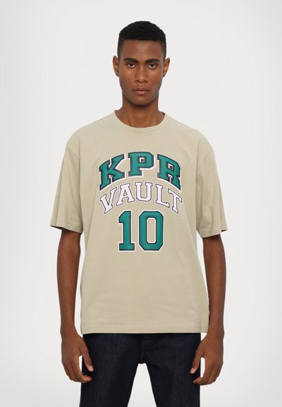 Bežs kokvilnas t-krekls ar lielu tumši zaļu un balto grafiku "KPR VAULT 10" uz priekšpuses. Īsās piedurknes un brīvs griezums.