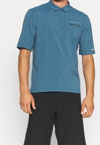 Homme portant un polo à manches courtes bleu avec des boutons et un pantalon noir, se tenant devant un fond clair.