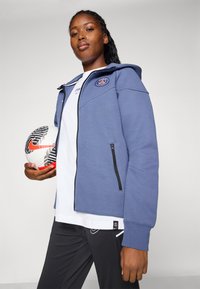 Blå hettegenser med glidelås og logo, lagd over en hvit skjorte, kombinert med svarte bukser. Holder en mønstrete fotball.