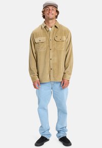 Tannenkleurige corduroy shirt met twee borstzakken, knoopsluiting en kraag. Gekleed met lichtblauwe jeans en zwarte schoenen.