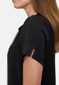 Svart T-shirt med korta ärmar, med slitsar och metallnitar vid ärmsluten. Mjuk textur, avslappnad passform och minimalistisk design.