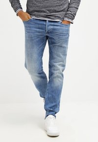 En person klädd i blå tvättade jeanjackor, grå långärmad tröja, händerna i fickorna och vita sneakers, går mot en enkel vit bakgrund.