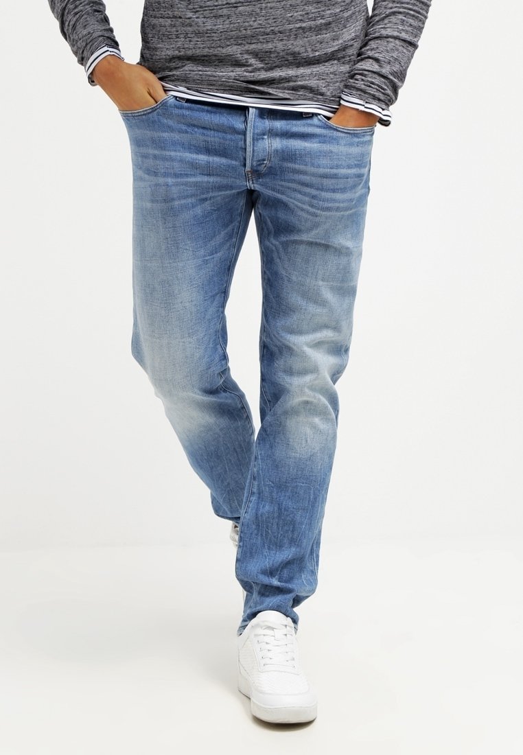 En person klädd i blå tvättade jeanjackor, grå långärmad tröja, händerna i fickorna och vita sneakers, går mot en enkel vit bakgrund.