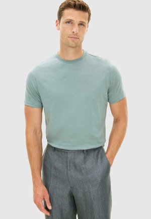 Mies, jolla on vaaleanvihreä lyhythihainen pyöreämpäinen paita ja harmaat juhlahousut, toinen käsi taskussa, yksinkertaisella taustalla.