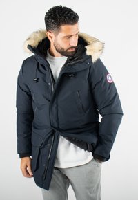 Veste matelassée marine avec capuche en fourrure, fermeture éclair et boutons, plusieurs poches et poignets élastiques. Équipée d'un patch logo sur la manche.