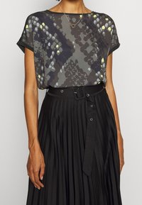 Krátký rukáv, šedý top s živými žlutými a modrými vzory; kombinovaný s skládací černou sukní s opaskem v pase a stříbrnými kovovými detaily.