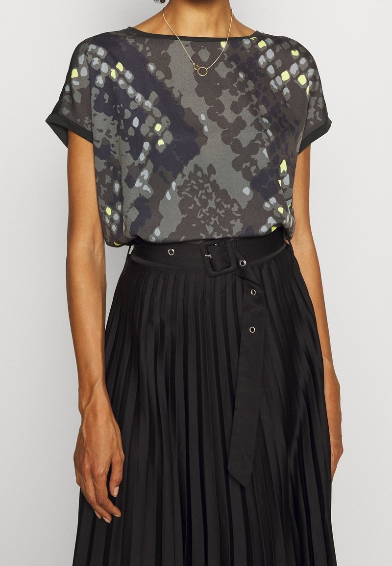 Krátký rukáv, šedý top s živými žlutými a modrými vzory; kombinovaný s skládací černou sukní s opaskem v pase a stříbrnými kovovými detaily.