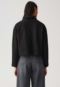 Persona con lunghi capelli scuri che indossa una giacca nera cropped e jeans neri sbiaditi a vita alta, vista di spalle su uno sfondo neutro.