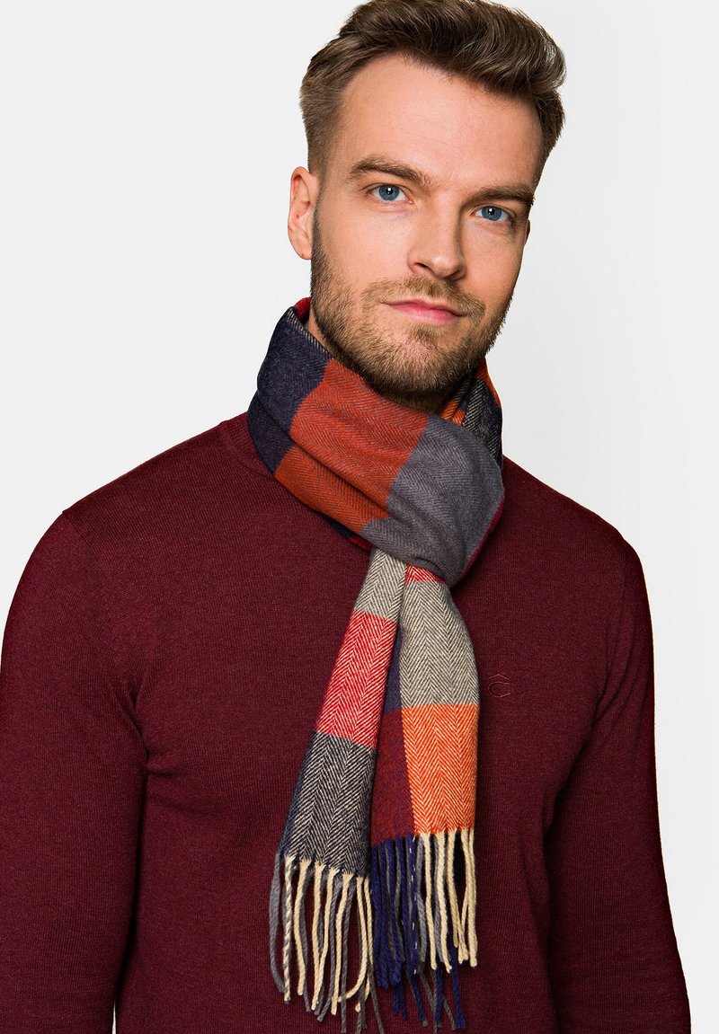 Szara, pomarańczowa i granatowa szalik w kratę z frędzlami. Luźno noszony na szyi na ciemnoczerwonym swetrze. Miękka faktura i wzór w jodełkę.