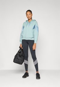 Lyseblå hettejakke med grå og blå paneler, parret med svarte leggings med grå striper og svarte treningssko. Bærer en svart treningsbag.