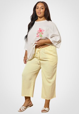 Frau steht vor einfarbigem Hintergrund und trägt ein übergroßes weißes T-Shirt mit Grafik, blassgelbe Hosen mit Kordelzug und weiße Slide-Sandalen.