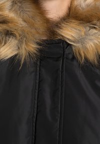 Manteau noir matelassé avec un col en fausse fourrure tan-brun duveteux, texture matelassée et fermeture éclair à l'avant.