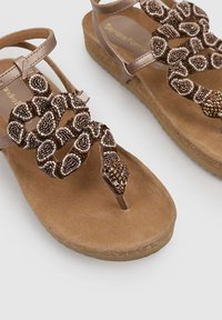Sandales plates perlées dans des tons bruns, présentant un accent de design en serpent avec des détails scintillants, sur une semelle légèrement texturée de couleur brune.