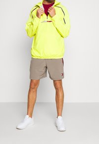 Neon gul vindjakke med halvlukket krave og sorte detaljer, parret med beige shorts med et logo patch og hvide sneakers.
