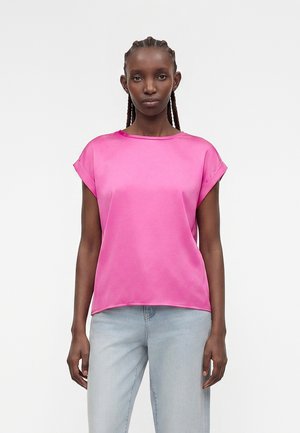 Γυναίκα με πλεξούδες στα μαλλιά, που φοράει μια φωτεινή ροζ μπλούζα με κοντά μανίκια και ανοιχτό μπλε τζιν, στέκεται μπροστά σε ένα ομοιόμορφο ανοιχτό φόντο.