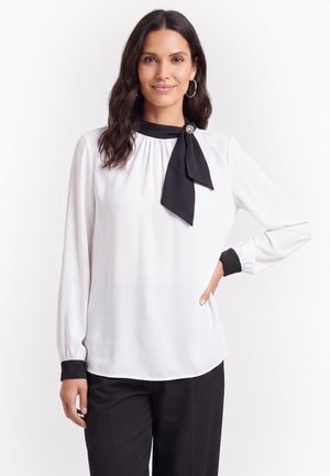 Vrouw met lang donker haar draagt een witte blouse met zwarte manchetten en stropdas, gecombineerd met een zwarte broek, glimlacht met één hand op de heup.