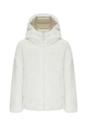 Jeune fille aux longs cheveux blonds portant une veste blanche gonflée, une jupe beige, des bottes noires et des chaussettes crème, debout devant un fond uni.