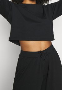 Sudadera negra corta con escote ancho y corte holgado, combinada con pantalones negros de cintura elástica a juego que cuentan con un cordón ajustable.