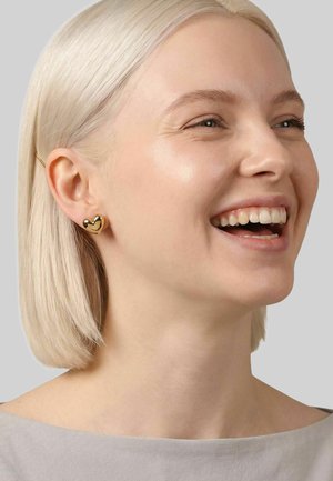 Vrouw met kort blond haar die glimlacht, draagt glanzende gouden oorstekers in de vorm van een hart, een lichtgrijze top, tegen een effen grijze achtergrond.