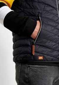 Main dans la poche zippée d'une veste matelassée bleu foncé portée sur une chemise à manches longues noire, blanche et jaune et un jean gris.