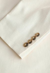 Manche de veste couleur crème avec quatre boutons marron cousus en rangée près du poignet sur un fond de tissu assorti.