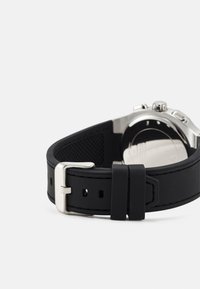 Orologio con cinturino in gomma nera e fibbia in metallo argento; presenta una finitura testurizzata e fori regolabili. Cassa in acciaio inossidabile visibile sul retro.