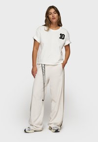 Witte gecropte t-shirt met zwart nummer "10" detail. Crème fleece broek met zijstrepen en een trekkoord. Gekocht met grijze en zwarte sneakers.