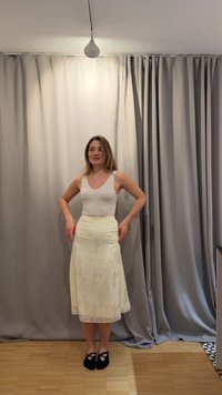 Γυναίκα στέκεται σε εσωτερικό χώρο σε ξύλινο πάτωμα, φοράει λευκό αμάνικο τοπ, κρεμ μίντι φούστα και μαύρες μπαλαρίνες, με τα χέρια κοντά στους γοφούς.