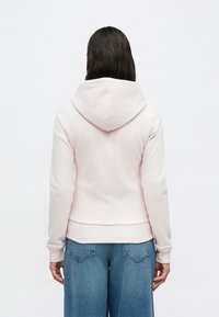 Ανοιχτό ροζ φούτερ με φαρδιά κουκούλα, ελαστικές μανσέτες και ριμπ τελείωμα. Το ύφασμα φαίνεται μαλακό και υφασμένο, με απλό σχεδιασμό.