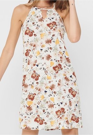 Femme portant une robe blanche sans manches avec un col halter et un imprimé floral multicolore dans des tons de brun, jaune et vert.