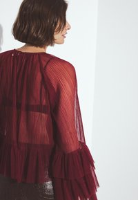 Blusa plissettata borgogna in tessuto trasparente, con maniche a volant e dettaglio chiave sul retro. Accentuata da una gonna aderente e lucida.