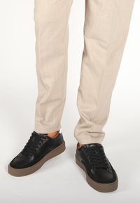 Scarpe da ginnastica in pelle nera con suola gommata marrone, dotate di lacci e un accento bianco a contrasto nella parte del tallone, indossate con pantaloni beige.