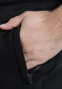 Schwarze Trainingshose mit einer Reißverschlusstasche an der Seite. Der Stoff wirkt glatt mit leichtem Stretch. Eine Hand ist in die Tasche gesteckt.