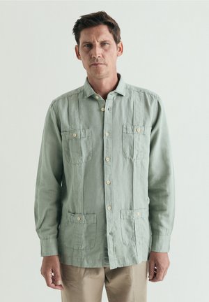 Hombre con expresión seria que lleva una camisa verde claro con botones y cuatro bolsillos, de pie frente a un fondo claro y liso.