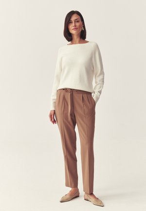 Kvinde med kort mørkt hår iført en hvid sweater, brune bukser med høj talje og bindebælte samt beige flade sko, stående foran en ensfarvet baggrund.